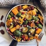 Crispy brussels sprouts with roasted apple and pecans topped with pomegranate seeds in a pot on a wood counter. There is a kitchen towel in the background and pecans.