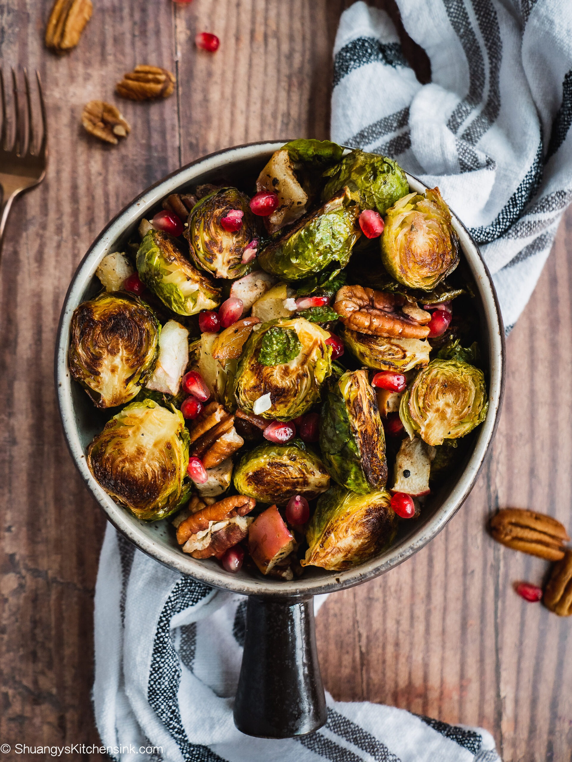 Crispy brussels sprouts with roasted apple and pecans topped with pomegranate seeds in a pot on a wood counter. There is a kitchen towel in the background and pecans.