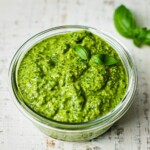 A dish of Vegan nut free pesto. There are a few pieces of basil on the side.