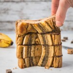 A stack of 4 pieces of banana bread with two pieces of sliced bananas on top. There is a hand grabbing one.
