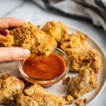 A plate of crispy and sizzling hot air fryer chicken nuggets. There is a serving dish of chick-fil-a copycat sauce in the middle. There is a hand holding a piece of chicken nugget.