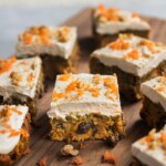 A freshly baked carrot cake cut into squares topped with cashew cream cheese frosting and sprinkle of fresh carrots, and walnuts.