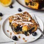 A bite shot is taken from a piece of healthy breakfast dessert. There is a crumbly top and a drizzle of maple coconut butter. There is also blueberries and lemons in the picture