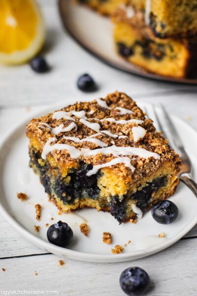 A bite shot is taken from a piece of healthy breakfast dessert. There is a crumbly top and a drizzle of maple coconut butter. There is also blueberries and lemons in the picture