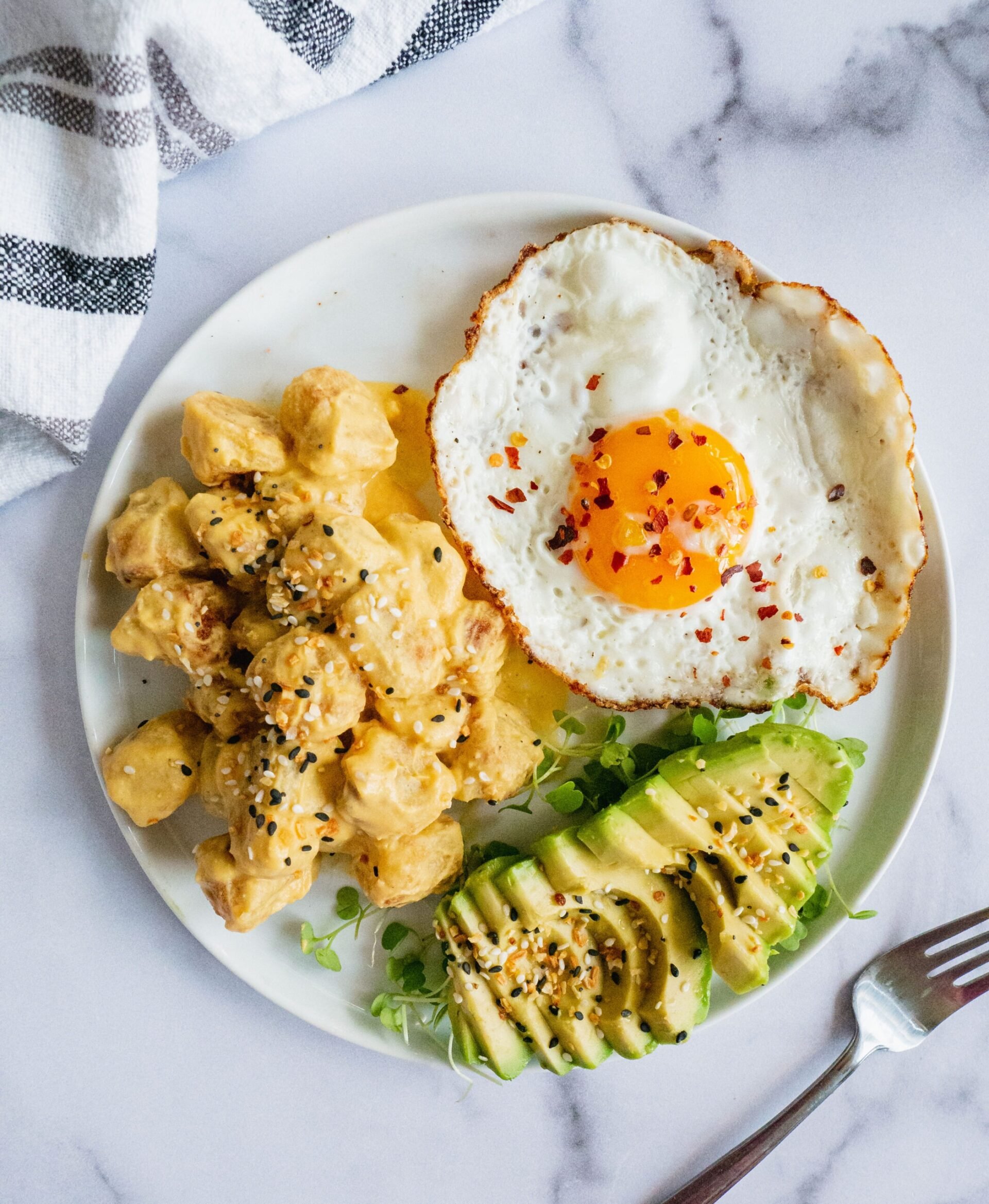 A plate of breakfast food. There is a fried egg with chili flakes, a sliced avocado and vegan cheese sauce