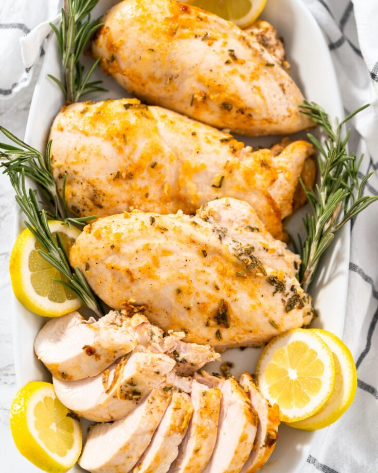 A serving dish with juicy chicken breast. There are lemon and Rosemary as well