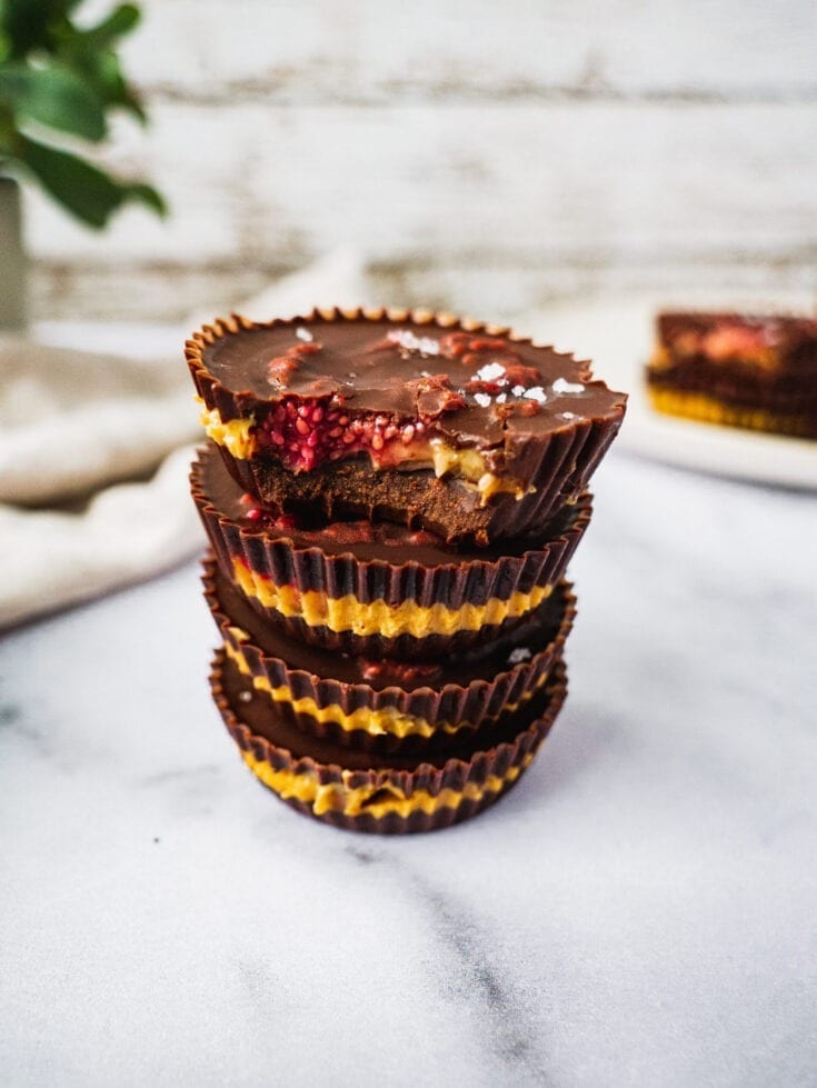 4 peanut butter jelly chocolate cups stack on top of each other. The top cup has a bite. Inside there is the raspberry chia jam and peanut butter banana filling, with sprinkle of sea salt on top.