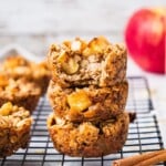 a Stack of Baked Apple Oatmeal Bake cups with an apple in the background and a cinnamon stick in the foreground.