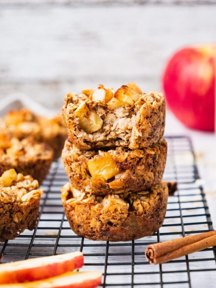 a Stack of Baked Apple Oatmeal Bake cups with an apple in the background and a cinnamon stick in the foreground.
