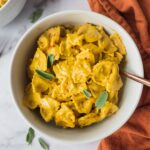A bowl of creamy and healthy pumpkin alfredo pasta on an orange dish towel with a couple sprigs of sage on top.