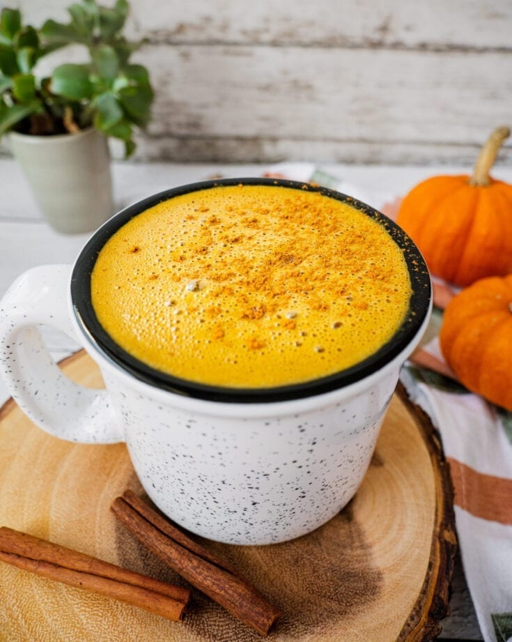A mug full of pumpkin spice latte with 2 pieces of cinnamon sticks in the foreground and 2 little pumpkins in the background.
