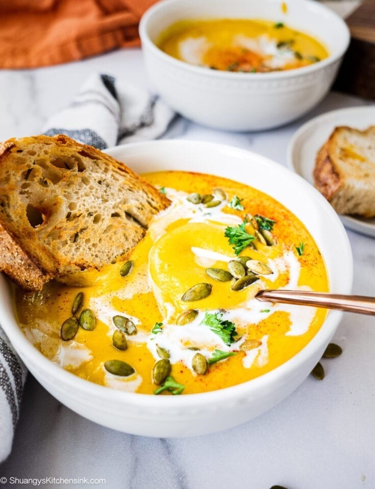 A Bowl Roasted Butternut Squash soup topped with coconut milk, toasted pumpkin seeds, parsley and a piece of crusty bread. There is another bowl in the background.