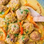 In a blue sauce pan, there are the cooked yellow thai coconut curry meatballs with fresh cilantro on top. There is a wooden spoon in the coconut curry soup as well.