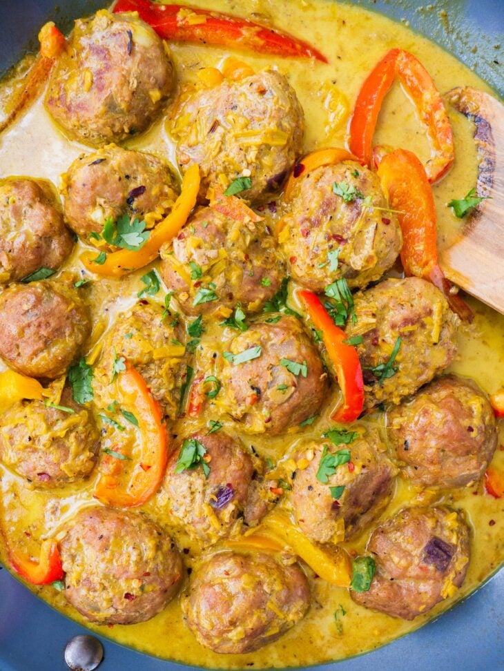 In a blue sauce pan, there are the cooked yellow thai coconut curry meatballs with fresh cilantro on top. There is a wooden spoon in the coconut curry soup as well.