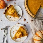 On a table there is a healthy pumpkin pie being sliced to serve. There are 3 little pumpkins around the pie. Two slices of pumpkin pies are placed on little plates, topped with whipped cream and pumpkin spice. There are two forks on the side.
