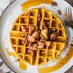 A paleo pumpkin waffle topped with sautéed apples and spiced pecans. Also drizzle of maple syrup. There is another waffle in the background and a little pumpkin on the kitchen towel.