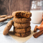 A stack of paleo ginger bread cookies with a glass of milk and cinnamon sticks in the background. The top cookie has a bite in it. The texture is chewy and soft.
