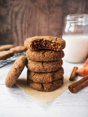 A stack of paleo ginger bread cookies with a glass of milk and cinnamon sticks in the background. The top cookie has a bite in it. The texture is chewy and soft.