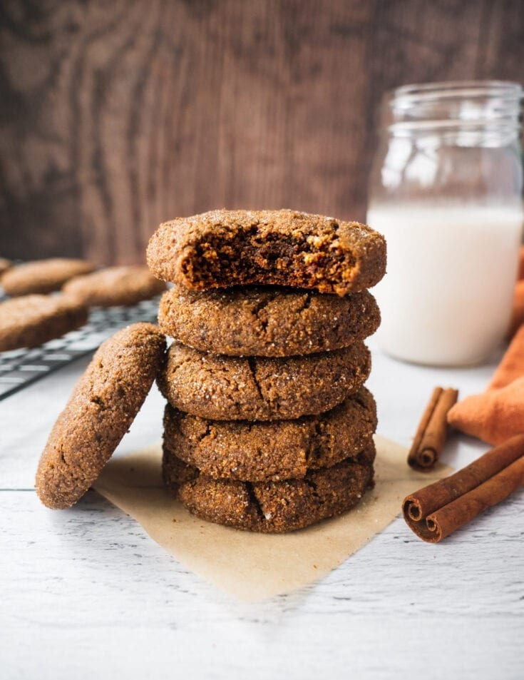 A stack of paleo ginger bread cookies with a glass of milk and cinnamon sticks in the background. The top cookie has a bite in it. The texture is chewy and soft.