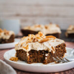 A piece of vegan gingerbread cake with cashew cream frosting and crumbled pecans on top. There is bite in the middle and a fork on the side. There are more gingerbread cake in the background.