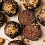 A batch of vegan chocolate truffles that is gingerbread flavored, topped with gingersnap cookies. One of the chocolate truffle is cut in half and the texture is gooey and smooth.