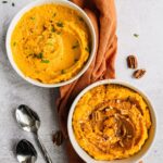 Two bowls of Sweet Potato Cauliflower Mash. One is maple pecan cinnamon sweet flavor, the other one is olive oil chive savory flavor. There are two spoons and an orange towel in the background as well.