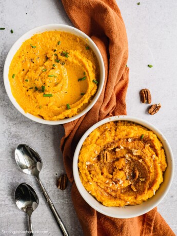 Two bowls of Sweet Potato Cauliflower Mash. One is maple pecan cinnamon sweet flavor, the other one is olive oil chive savory flavor. There are two spoons and an orange towel in the background as well.