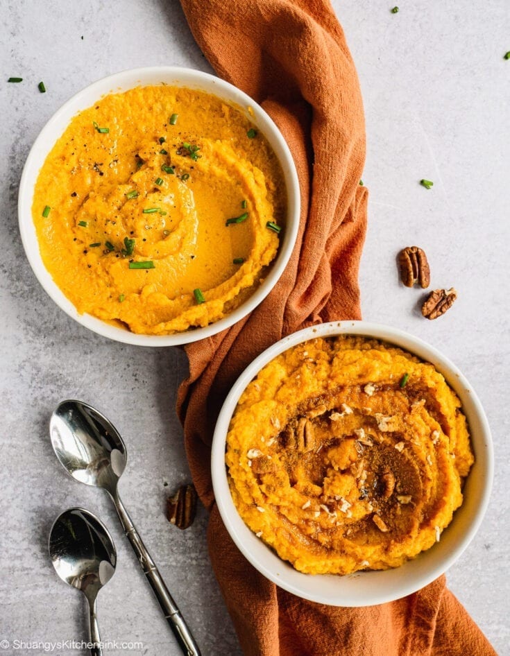 Two bowls of Sweet Potato Cauliflower Mash. One is maple pecan cinnamon sweet flavor, the other one is olive oil chive savory flavor. There are two spoons and an orange towel in the background as well.
