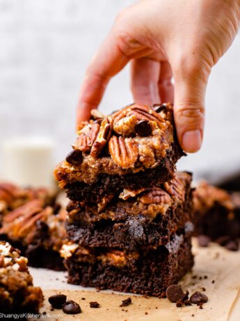 A hand is reaching for a stack of ooey gooey pecan pie brownies topped with whole nuts and dark dairy free chocolate chips