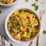 A bowl of cauliflower fried rice with chicken that is colorful and so flavorful. There is a pair of chopsticks and some fresh cilantro on the table.