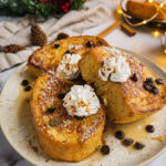 A plate of dairy free eggnog french toast topped with dairy free whip cream and chocolate chips. There is Christmas decoration in the background.