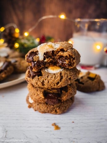 A stack of chocolate chip smores cookies. The top cookie has a bite in it. There are Christmas lights, and a glass of milk in the background.