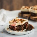 A piece of chocolate cake with cream cheese frosting with a bite in the middle. There are pecans on top.