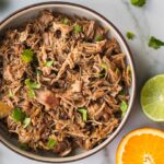 A bowl of pulled pork carnitas with a few pieces of cilantro on top. And there is half of a piece of orange in the back.