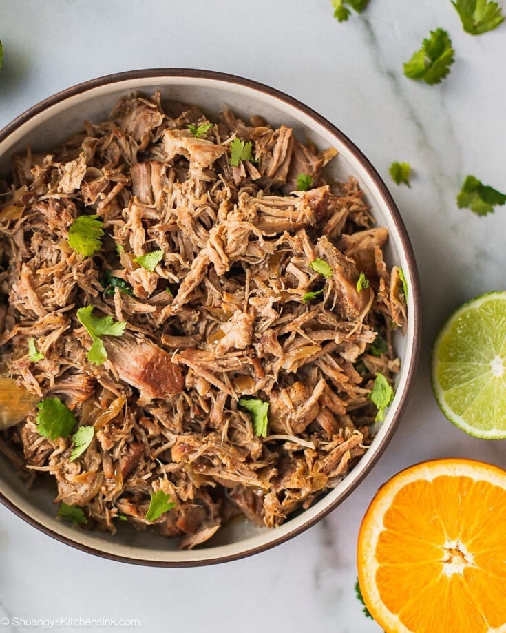 A bowl of pulled pork carnitas with a few pieces of cilantro on top. And there is half of a piece of orange in the back.