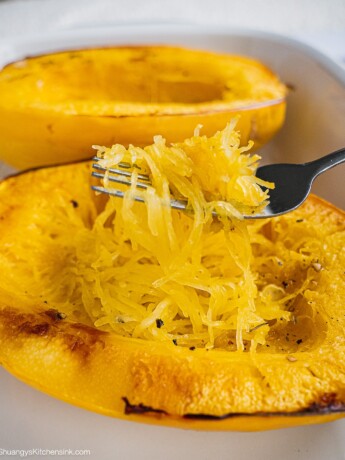 Two baked Spaghetti Squash on a baking dish. There is a fork shredding Spaghetti Squash into strands.