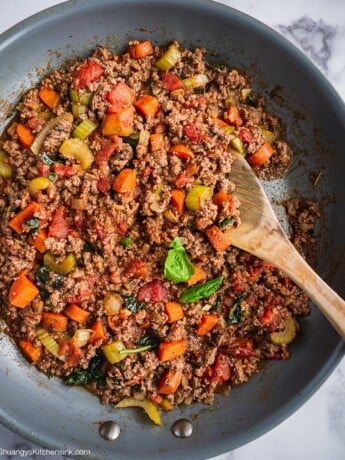 A pan of fresh and cooked bolognese sauce. There are carrots, celery and ground meat. There are some fresh basil on top.