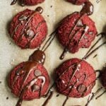 A parchment paper lined pan with red velvet edible cookie dough on top. There are chocolate chips and melted chocolate drizzle on top.