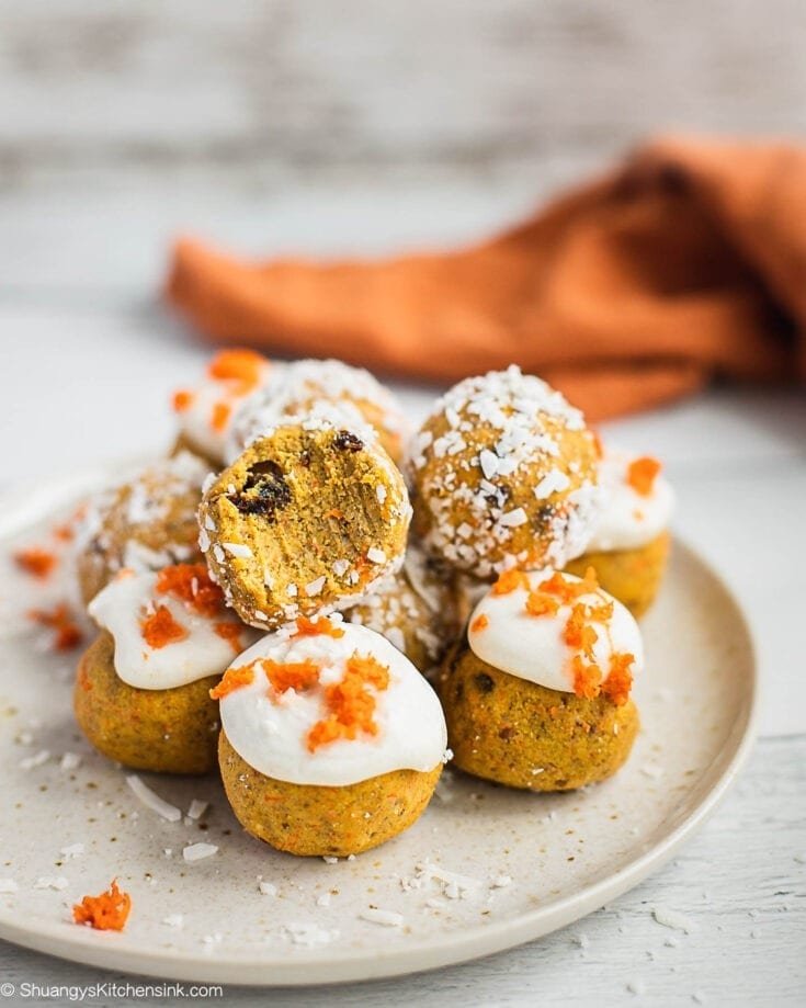 A plate of Carrot Cake truffles. A few of them are covered by coconut flakes, a few are covered by coconut butter and topped with shredded carrots. There is a bite on top.