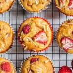 A batch of freshly baked strawberry muffins on a cooling rack. There are a few pieces of fresh strawberries on the side.
