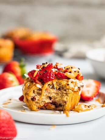 There is a strawberry banana muffin on a plate, topped with nut butter, fresh strawberry. There is a bite.