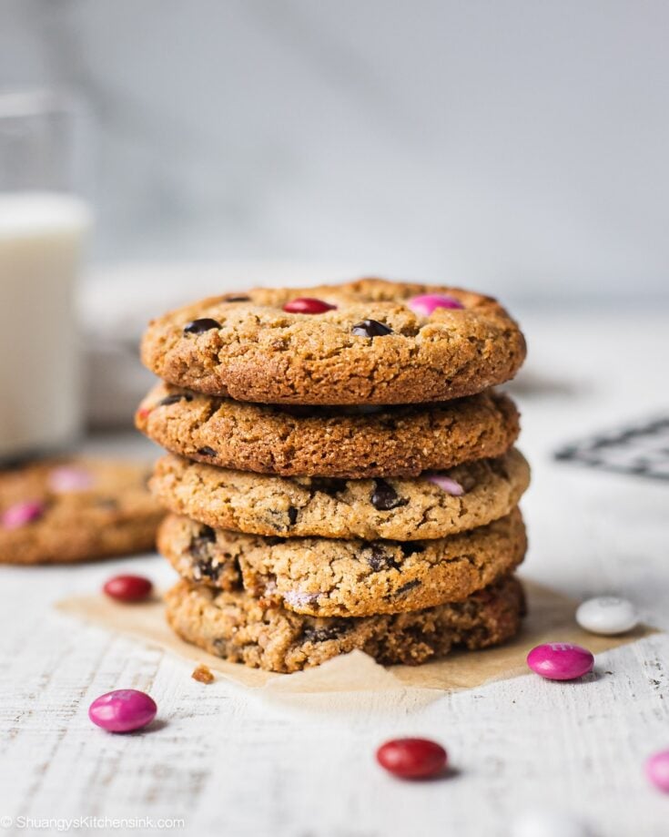 A stack of eggless chocolate chip cookies with M&M that can be made keto, and paleo as well. Perfect for a healthy Valentine's Day dessert. There are a few pink M&Ms on the table and a glass of milk in the background to serve the cookies with.
