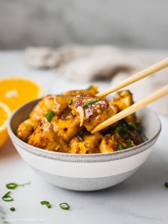 A bowl fo whole30 orange chicken ready to be served. The healthy orange chicken is juicy and tender, coated with tangy orange sauce. There is a pair of chopsticks.