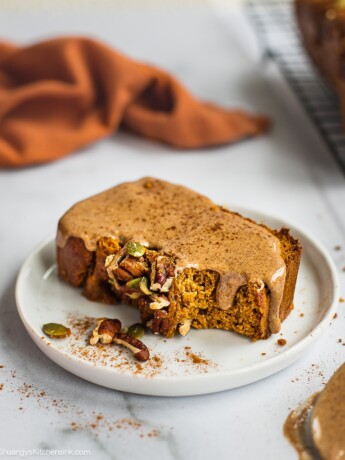 A slice of paleo sweet potato bread on a plate. It is topped with nut butter and cinnamon. there is a cooling rack and the rest of the loaf in the back.