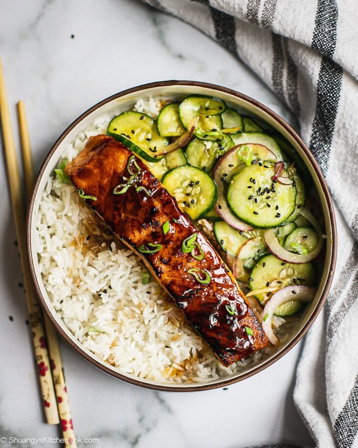 A bowl of jasmine rice crispy teriyaki salmon, asian cucumber salad. There is a pair of chopsticks on the side.