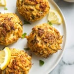 A plate of golden brown and crispy crab cakes on a plate. There are some lemon and chopped parsley for serving.