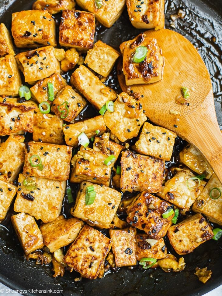 Golden brown cubes of tofu cooked in a skillet with sauce, inspired by classic Chinese tofu recipes, and garnished with sliced green onions and black sesame seeds. A wooden spoon stirs the tofu pieces.