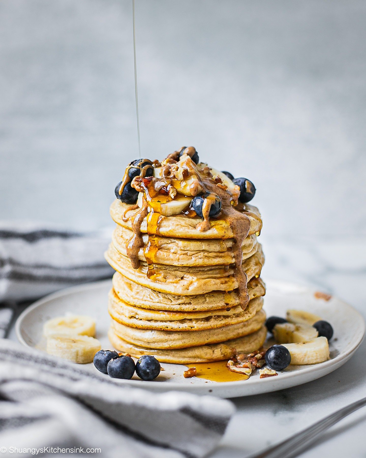 A stack of healthy buttermilk pancakes topped with bananas, blueberries, almond butter, and pecans. There is maple syrup dripping down from the top.