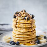 A stack of healthy buttermilk pancakes topped with bananas, blueberries, almond butter, and pecans. There is maple syrup dripping down from the top.