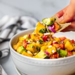 A bowl of fresh ingredients for mango avocado salsa that include fresh mango, avocado, red onion, tomatoes, diced jalapeno, and cilantro. There are chips on the side. A hand is digging in with some chips.